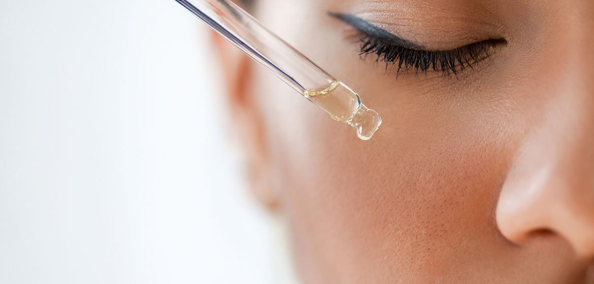 Nahaufnahme der rechten Gesichtshälfte einer jungen Frau. Sie hat ihr mit Mascara und Kajal geschminktes Auge geschlossen. Eine Pipette mit einer klaren Flüssigkeit nähert sich ihrer Wange.